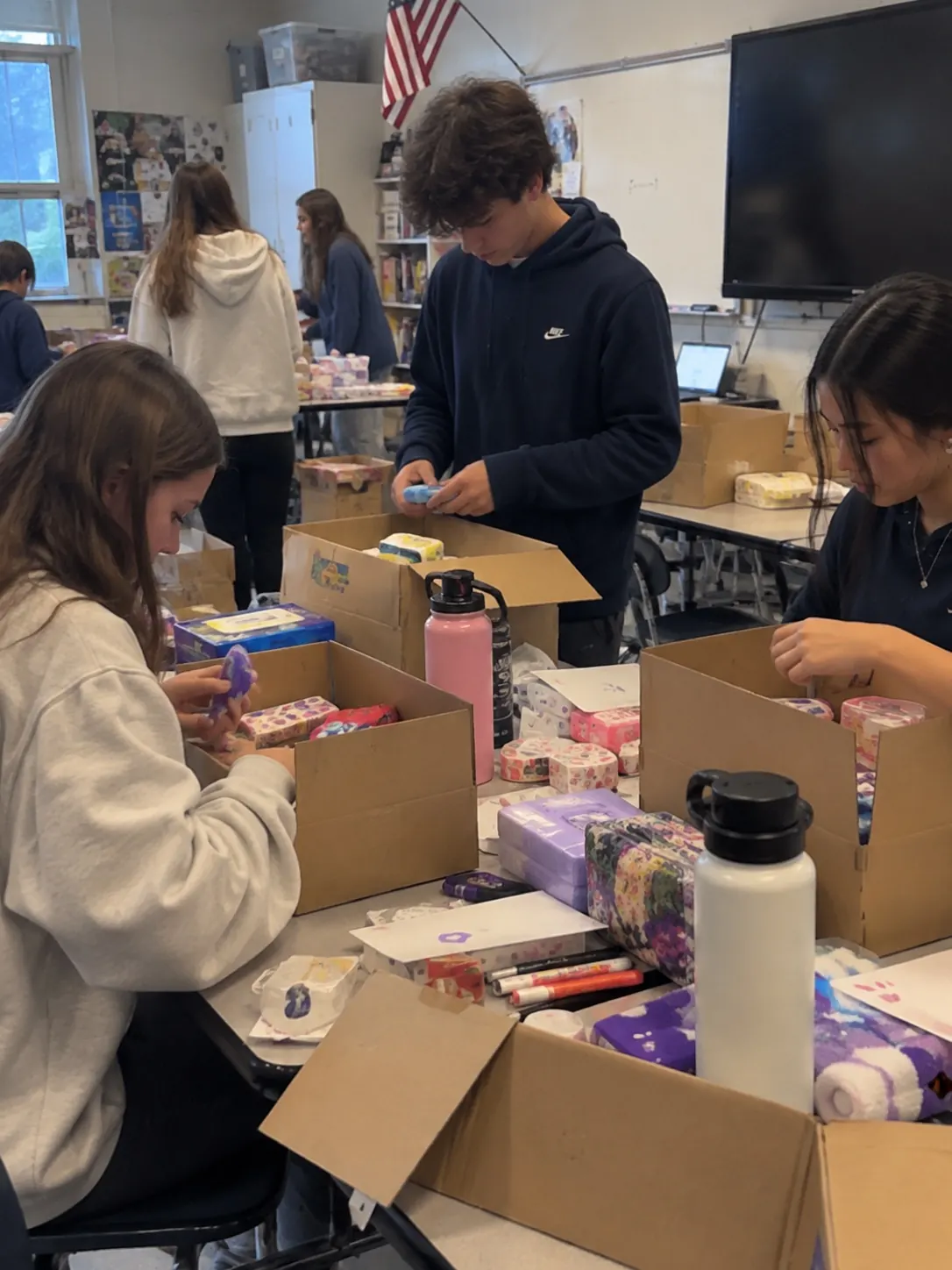 Detailing for MCAS student volunteers packing care packages into boxes in a classroom