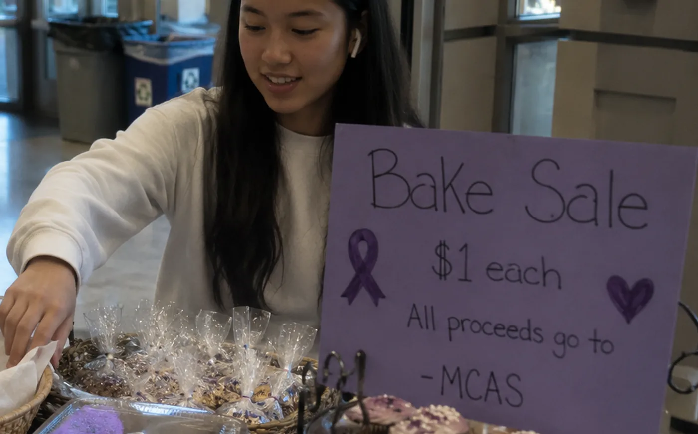 A student volunteer setting out baked goods beside a hand-lettered sign reading &ldquo;Bake Sale, $1 each, all proceeds go to MCAS&rdquo;