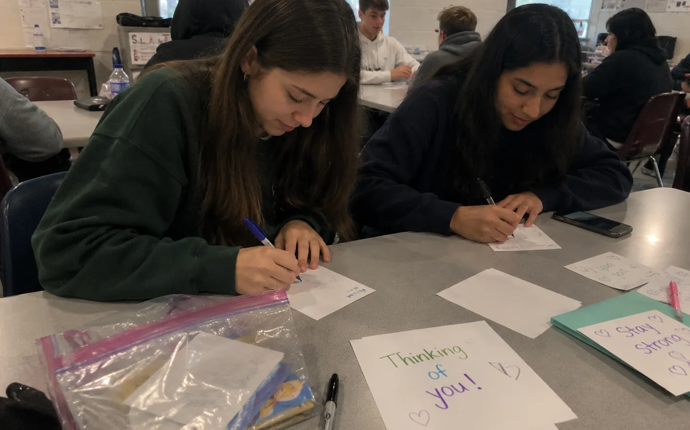 Two student volunteers writing inspiration cards at a classroom table, one card reading &ldquo;Thinking of you&rdquo;